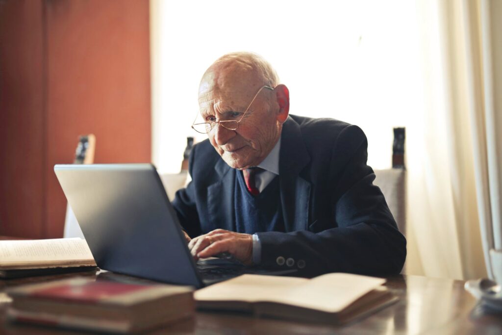 Concentrate elderly senior man in formal suit and eyeglasses working on laptop while sitting at wooden table with books in light room